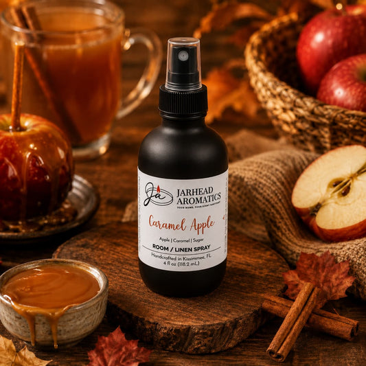 A bottle of Jarhead Aromatics Caramel Apple Room & Linen Spray | Crisp Apple, Caramel & Warm Spice rests on a table surrounded by caramel, apples, cinnamon sticks, and a mug in the background, creating a cozy autumn ambiance and infusing the air with the perfect fall home fragrance.