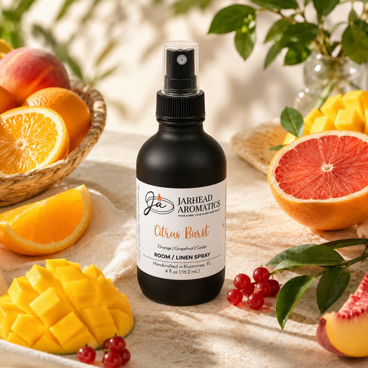 A bottle of Jarhead Aromatics Citrus Burst Room & Linen Spray | Orange, Mango, Grapefruit & Peach rests on a table surrounded by orange slices, grapefruit halves, peaches, and red berries, while leafy green branches provide a lush backdrop. This home fragrance features a lively blend of tropical fruits to instantly brighten any space.
