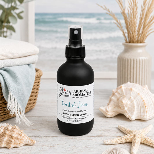 A black spray bottle labeled Jarhead Aromatics Coastal Linen Room & Linen Spray | Fresh Linen, Ozone & Sandalwood rests on a white table, surrounded by seashells, towels, a woven basket, and beach scenery in the background—evoking a refreshing home fragrance perfect for any space.