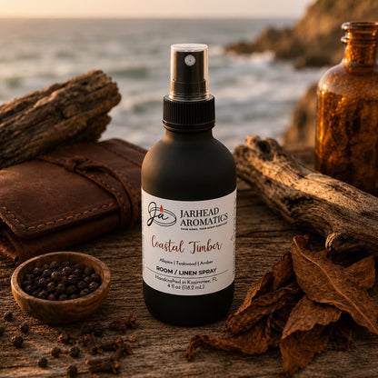 A bottle of Jarhead Aromatics Coastal Timber Room & Linen Spray | Teakwood, Leather & Warm Amber rests on driftwood near dried leaves, peppercorns, a brown glass bottle, and the ocean at sunset—embodying the refined essence of this luxury home fragrance with its evocative woodsy notes.