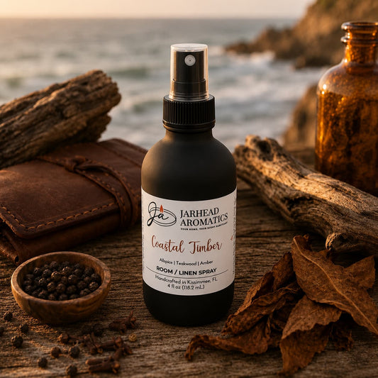 A bottle of Jarhead Aromatics Coastal Timber Room & Linen Spray | Teakwood, Leather & Warm Amber rests on driftwood near dried leaves, peppercorns, a brown glass bottle, and the ocean at sunset—embodying the refined essence of this luxury home fragrance with its evocative woodsy notes.