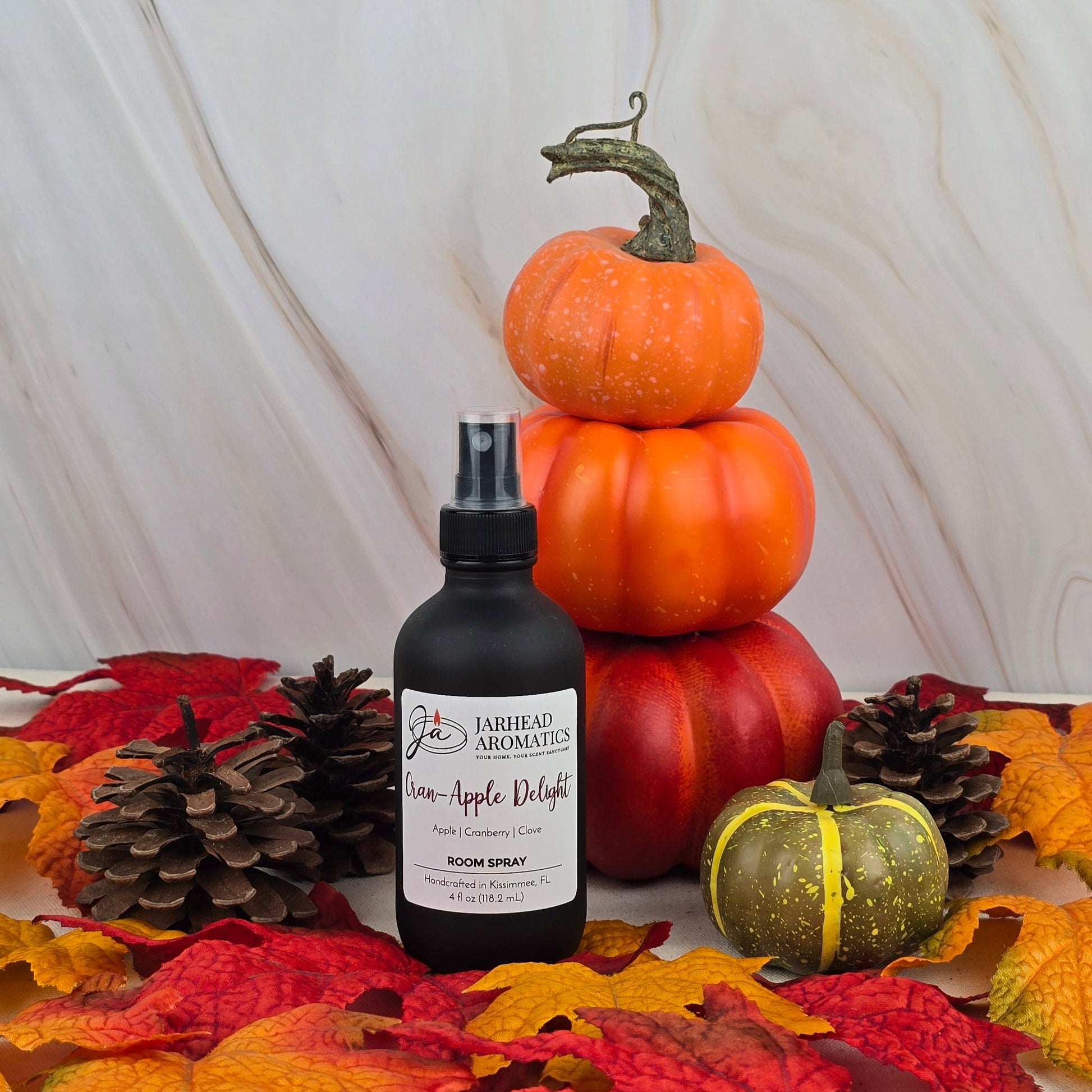A bottle of Jarhead Aromatics Cran-Apple Delight Room Spray from the Fall Collection sits among mini pumpkins, pinecones, and autumn leaves, creating a cozy fall-themed display.