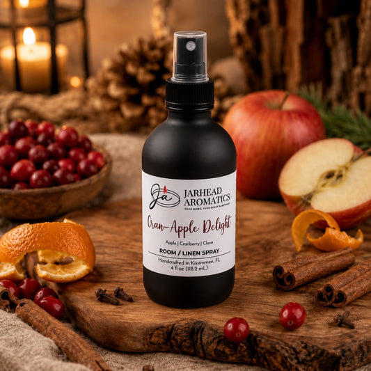 A bottle of Jarhead Aromatics Cran-Apple Delight Room & Linen Spray | Cranberry, Apple & Warm Cinnamon sits on a wooden board surrounded by apples, cranberries, orange peel, and cinnamon sticks, set against rustic decor. This inviting display beautifully reflects the cozy blend of fruit and warm spice that makes this room and linen spray a welcoming addition to any home.