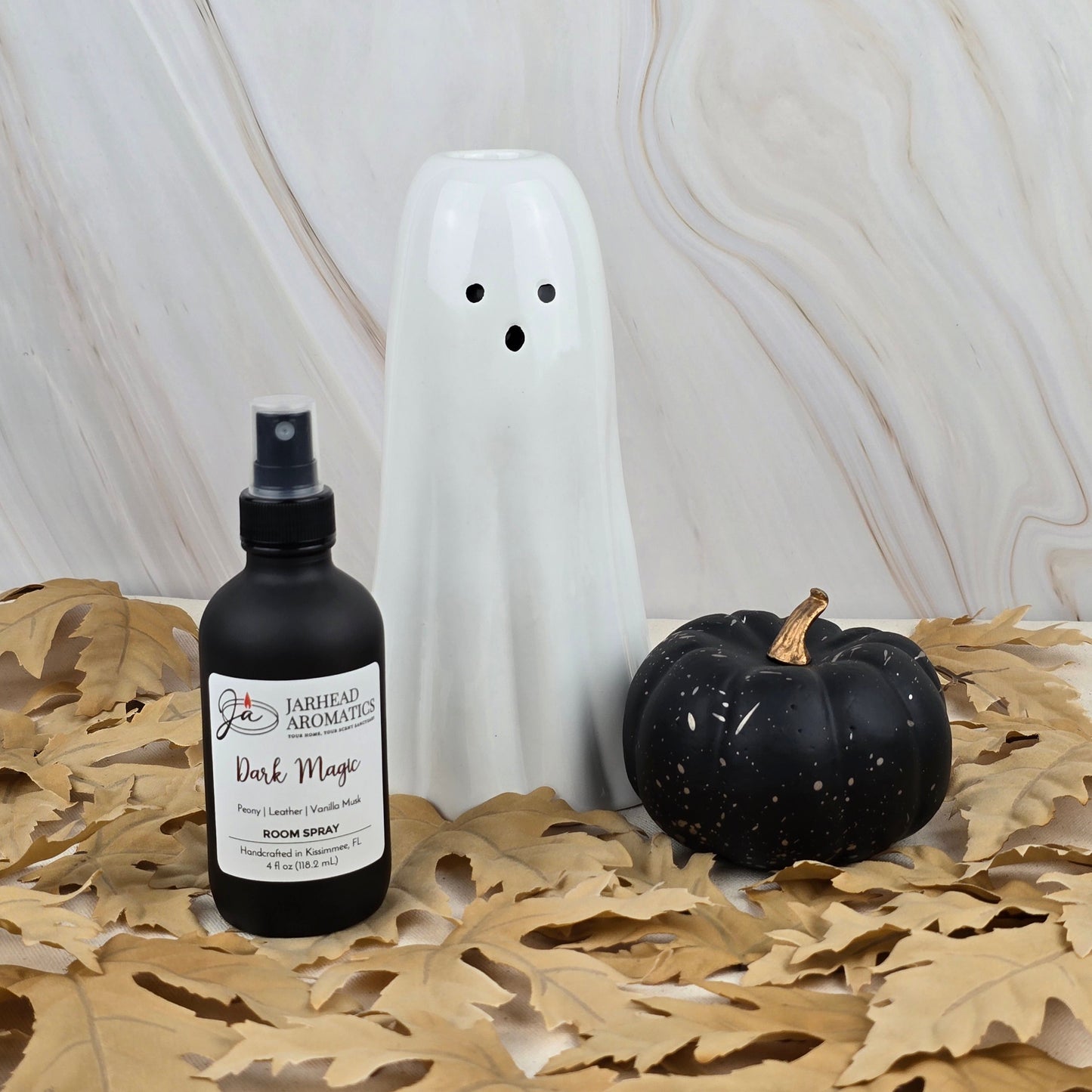 A ceramic ghost, a black pumpkin, and Jarhead Aromatics’ Dark Magic Room Spray from the Halloween Collection sit on dried leaves with a marble background, forming a spooky and fragrant seasonal display.
