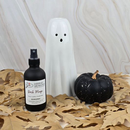 A ceramic ghost, a black pumpkin, and Jarhead Aromatics’ Dark Magic Room Spray from the Halloween Collection sit on dried leaves with a marble background, forming a spooky and fragrant seasonal display.