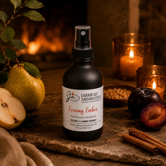 A bottle of Jarhead Aromatics Evening Ember Room & Linen Spray | Plum, Sandalwood & Soft Smoke rests on a cozy wooden surface surrounded by pears, plums, cinnamon sticks, a candle, and autumn leaves with a fireplace in the background. This inviting home fragrance blends juicy plum with warm sandalwood and soft smoke notes to create an atmosphere of comfort and relaxation.