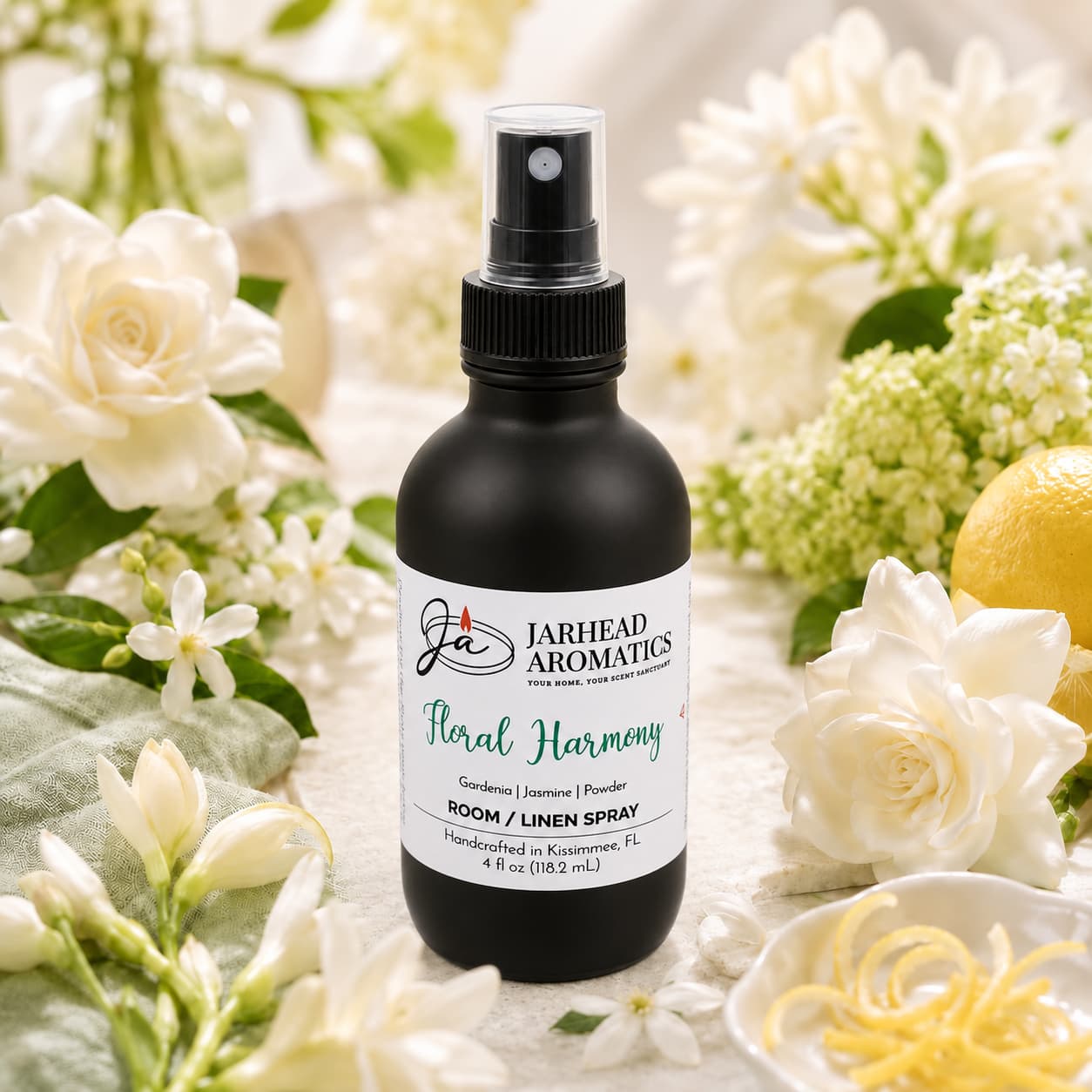 A black spray bottle labeled “Floral Harmony Room & Linen Spray | Gardenia, Jasmine & Tuberose” by Jarhead Aromatics rests among white flowers, green leaves, and a lemon on a light surface, creating a fresh and elegant scene that perfectly embodies the essence of home fragrance. This luxurious spray fills any space with bright, uplifting notes reminiscent of blooming gardenia, jasmine, tuberose, and citrus.