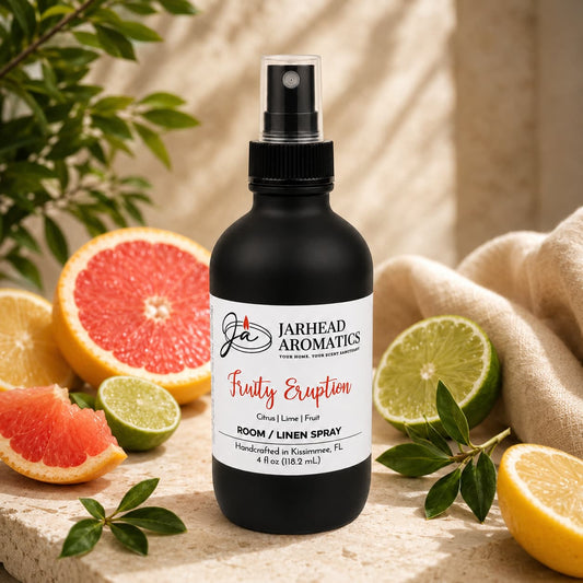 A sleek black spray bottle of Jarhead Aromatics’ Fruity Eruption Room & Linen Spray | Lemon, Lime, Grapefruit & Fresh Fruit sits on a stone surface surrounded by sliced grapefruit, lime, and orange, radiating the lively essence of a citrus home fragrance. Leafy branches and a beige textured cloth in the background enhance this inviting display.