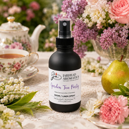 A bottle of Jarhead Aromatics Garden Tea Party Room & Linen Spray | Rose, Lilac & Jasmine rests on a floral tablecloth, surrounded by flowers, a teacup, a teapot, and a pear in the background. This inviting scene captures the refreshing ambiance of a garden tea party and highlights the floral home fragrance that fills your space with rose, lilac, and jasmine-inspired charm.