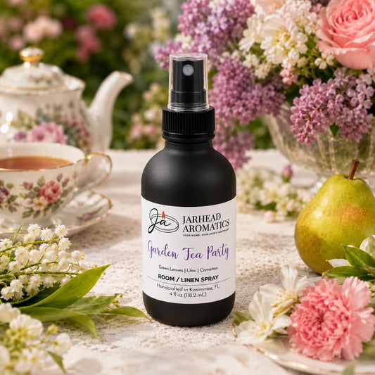 A bottle of Jarhead Aromatics Garden Tea Party Room & Linen Spray | Rose, Lilac & Jasmine rests on a floral tablecloth, surrounded by flowers, a teacup, a teapot, and a pear in the background. This inviting scene captures the refreshing ambiance of a garden tea party and highlights the floral home fragrance that fills your space with rose, lilac, and jasmine-inspired charm.