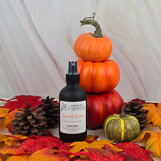 A bottle of Jarhead Aromatics Harvest Cream Room Spray from the Fall Collection sits among pinecones, faux autumn leaves, and stacked pumpkins, adding a cozy fall scent to the marble-backed festive display.