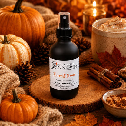 A bottle of Jarhead Aromatics Harvest Cream Room & Linen Spray—featuring notes of pumpkin spice, brown sugar, and vanilla cream—rests on a wooden slab amid pumpkins, autumn leaves, pinecones, cinnamon sticks, a cozy sweater, and a whipped cream-topped mug in a warm fall setting—evoking the ultimate gourmand home fragrance experience.