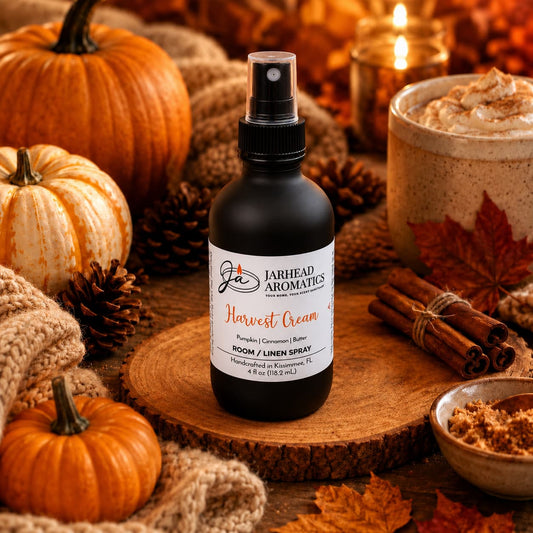 A bottle of Jarhead Aromatics Harvest Cream Room & Linen Spray—featuring notes of pumpkin spice, brown sugar, and vanilla cream—rests on a wooden slab amid pumpkins, autumn leaves, pinecones, cinnamon sticks, a cozy sweater, and a whipped cream-topped mug in a warm fall setting—evoking the ultimate gourmand home fragrance experience.