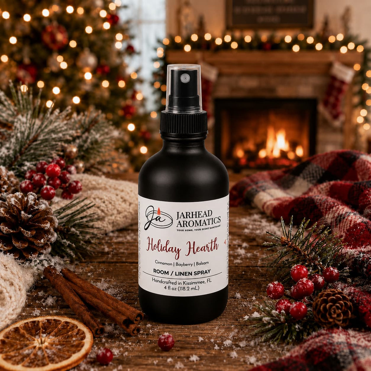 A black bottle of Jarhead Aromatics Holiday Hearth Room & Linen Spray sits on a wooden surface decorated with pine branches, cinnamon sticks, and dried orange slices.