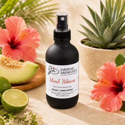 A bottle of Jarhead Aromatics Island Hibiscus Room & Linen Spray | Honeydew, Lime & Tropical Florals is displayed on a light stone surface, accompanied by vibrant hibiscus flowers, half a melon, a lime, and lush green succulents.