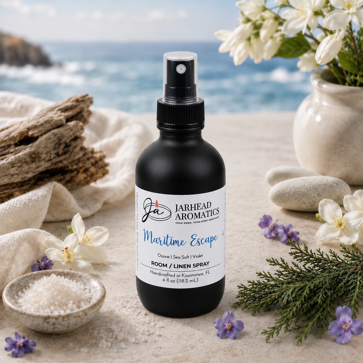 A bottle of Jarhead Aromatics Maritime Escape Room & Linen Spray | Sea Salt, Ozone & Jasmine rests on a table adorned with sea salt, flowers, and the ocean in the background, infusing your home with a fresh coastal scent for an elevated fragrance experience.