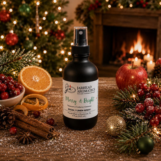 A black Jarhead Aromatics Merry & Bright Room & Linen Spray sits on a wooden surface surrounded by fresh cranberries, orange slices, cinnamon sticks, and pine branches in front of a fireplace.