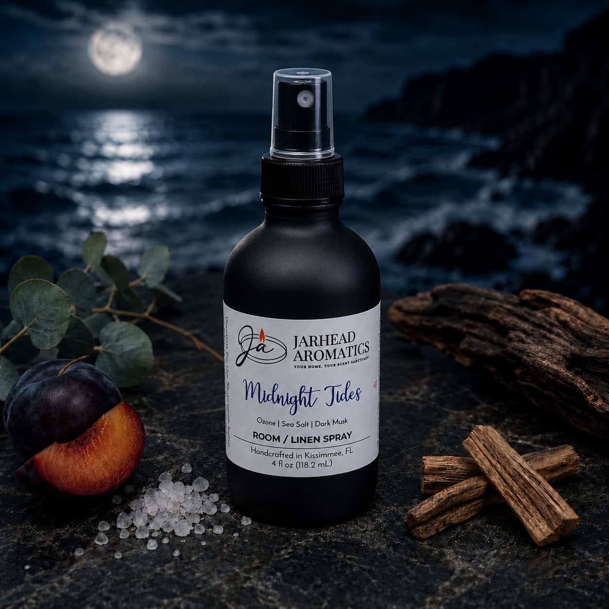 A bottle of Jarhead Aromatics’ Midnight Tides Room & Linen Spray—featuring notes of sea salt, sandalwood, and dark musk—rests on a dark surface with accents of plum, salt crystals, cinnamon sticks, wood, and eucalyptus that evoke its coastal fragrance. In the background, an ocean glimmers under a moonlit sky.