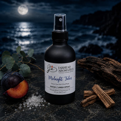 A bottle of Jarhead Aromatics’ Midnight Tides Room & Linen Spray—featuring notes of sea salt, sandalwood, and dark musk—rests on a dark surface with accents of plum, salt crystals, cinnamon sticks, wood, and eucalyptus that evoke its coastal fragrance. In the background, an ocean glimmers under a moonlit sky.