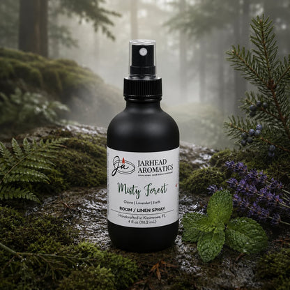 A black spray bottle labeled “Misty Forest Room & Linen Spray | Ozone, Moss & Juniper” by Jarhead Aromatics rests atop lush moss in a woodland scene, surrounded by verdant foliage and clusters of purple berries. Mist drifts through the trees in the background, evoking a nature-inspired home fragrance that captures the essence of a misty forest.