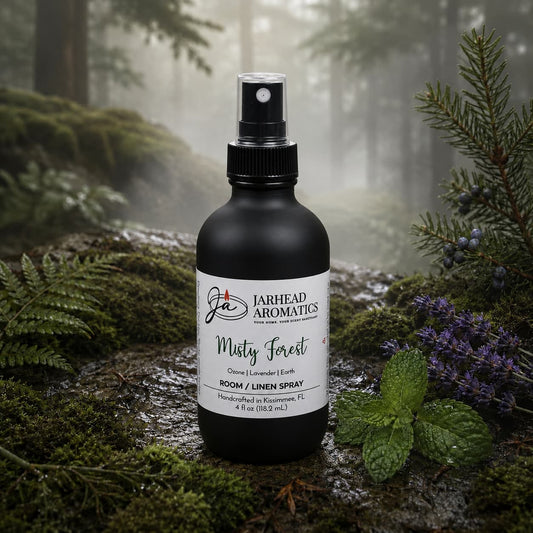 A black spray bottle labeled “Misty Forest Room & Linen Spray | Ozone, Moss & Juniper” by Jarhead Aromatics rests atop lush moss in a woodland scene, surrounded by verdant foliage and clusters of purple berries. Mist drifts through the trees in the background, evoking a nature-inspired home fragrance that captures the essence of a misty forest.