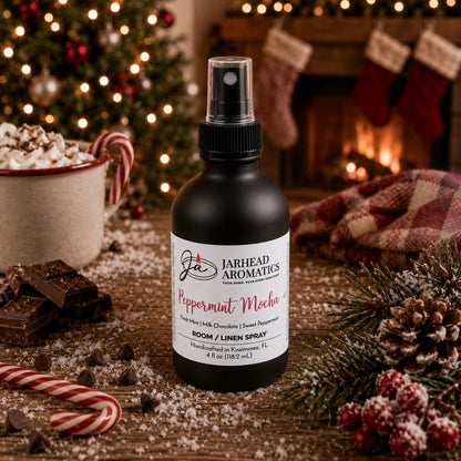 A bottle of Peppermint Mocha Room & Linen Spray | Chocolate Mint & Vanilla sits on a wooden table surrounded by chocolate pieces, a candy cane, and a mug of hot cocoa with a fireplace and Christmas tree in the background.