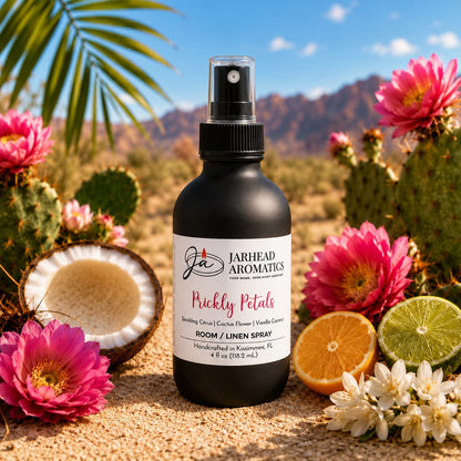 A bottle of Jarhead Aromatics Prickly Petals Room & Linen Spray | Cactus Flower, Coconut & Sandalwood rests on sunlit sand surrounded by vibrant pink cactus blooms, fresh citrus slices, and an open coconut, its desert oasis-inspired fragrance floating on the air with desert scenery and distant mountains in the background.