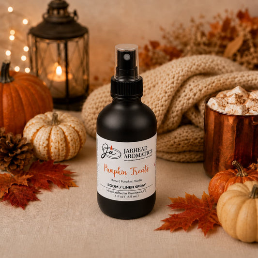 A bottle of Jarhead Aromatics’ Pumpkin Treats Room & Linen Spray—featuring notes of pumpkin, caramel, maple, and pecan—rests on a table surrounded by pumpkins, pinecones, cozy knit fabric, a lantern, and a mug of marshmallows, filling the space with an inviting cozy fall aroma that creates the perfect warm autumn atmosphere.