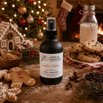 A black bottle of Santa’s Cookies Room & Linen Spray sits on a wooden table surrounded by gingerbread men, chocolate chip cookies, and spices in front of a fireplace.