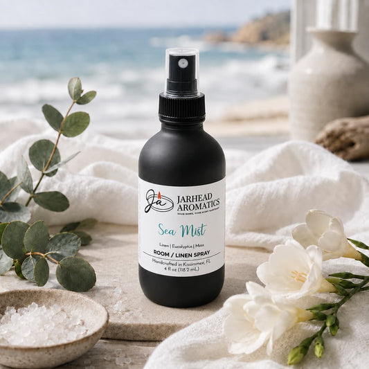 A black bottle of Jarhead Aromatics’ Sea Mist Room & Linen Spray—featuring notes of fresh linen, sea salt, and eucalyptus—rests on a table adorned with sea salt and white flowers, evoking the crisp essence of coastal home fragrance as a softly blurred beach and ocean create a serene backdrop.