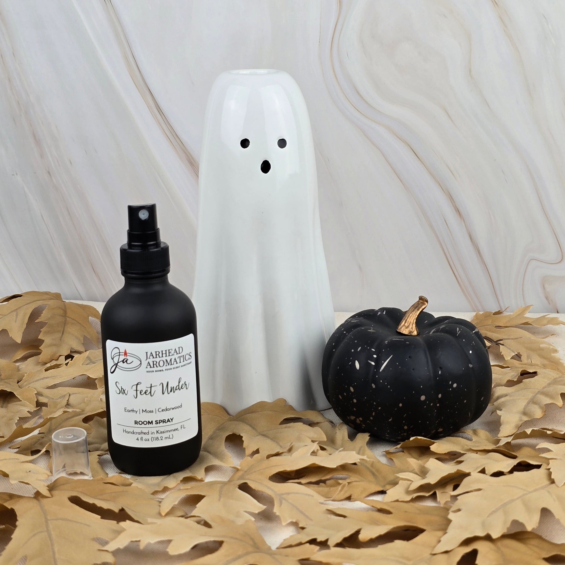 A ceramic ghost, a black pumpkin, and Jarhead Aromatics’ Six Feet Under Room Spray—part of the Halloween Collection and featuring earthy moss notes—are displayed on dried leaves with a marble backdrop.