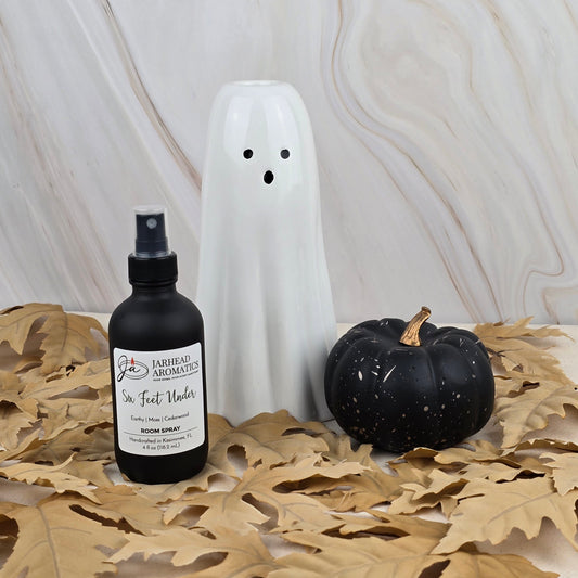 A ceramic ghost, a black pumpkin, and a bottle of Six Feet Under Room Spray by Jarhead Aromatics are arranged on dried oak leaves in front of a marbled background.