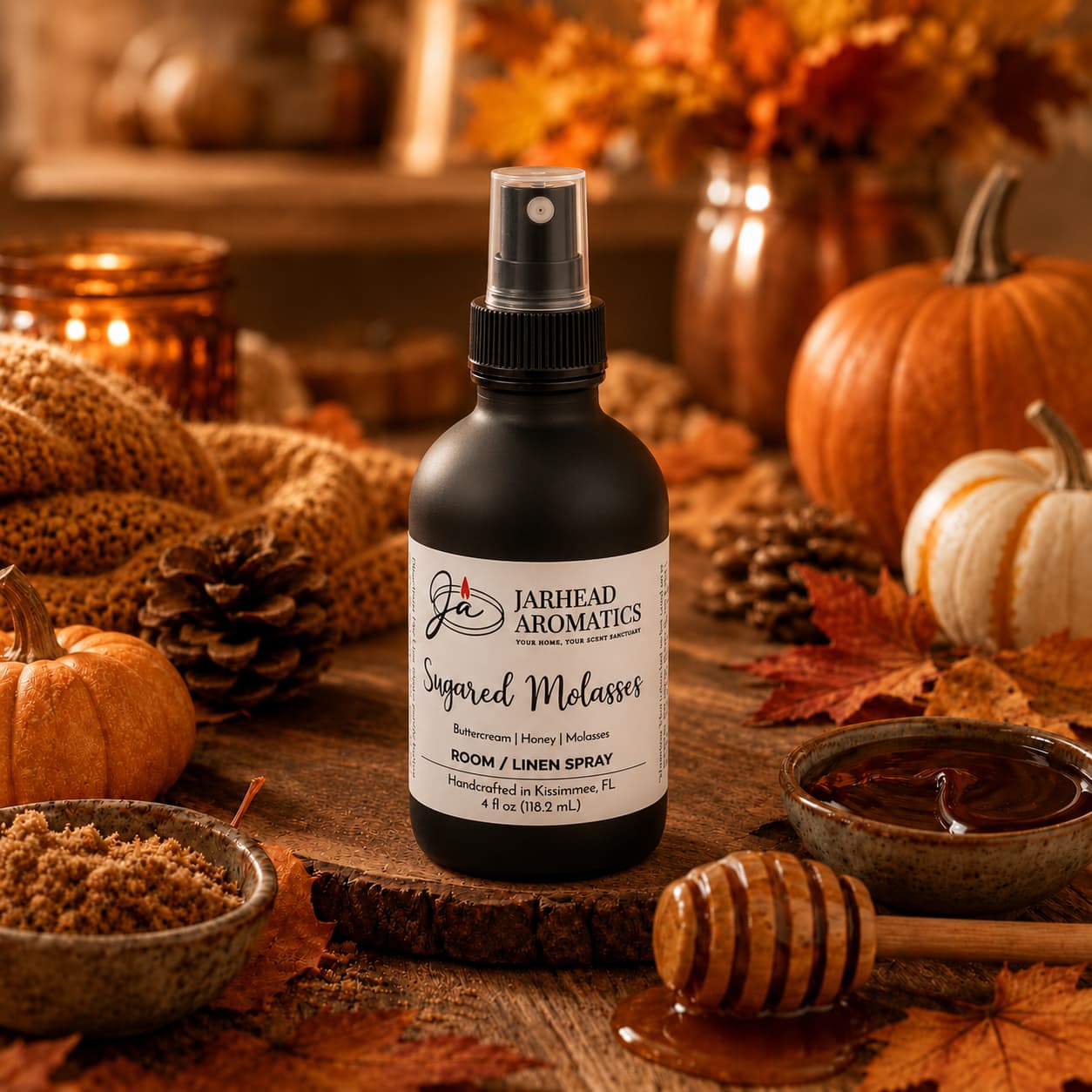 A bottle of Jarhead Aromatics Sugared Molasses Room & Linen Spray, featuring notes of buttercream, honey, and dark molasses, rests on a wooden table surrounded by autumn leaves, pumpkins, brown sugar, a honey dipper, and a bowl of syrup—creating a cozy fall atmosphere.