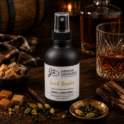A bottle of Jarhead Aromatics Sweet Bourbon Room & Linen Spray | Bourbon, Butterscotch & Brown Sugar is artfully arranged on a rustic table, surrounded by sugar cubes, cardamom pods, black peppercorns, a glass of bourbon, and a wooden barrel—evoking the warm and inviting gourmand notes that make this home fragrance truly unforgettable.