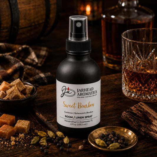 A bottle of Jarhead Aromatics Sweet Bourbon Room & Linen Spray | Bourbon, Butterscotch & Brown Sugar is artfully arranged on a rustic table, surrounded by sugar cubes, cardamom pods, black peppercorns, a glass of bourbon, and a wooden barrel—evoking the warm and inviting gourmand notes that make this home fragrance truly unforgettable.