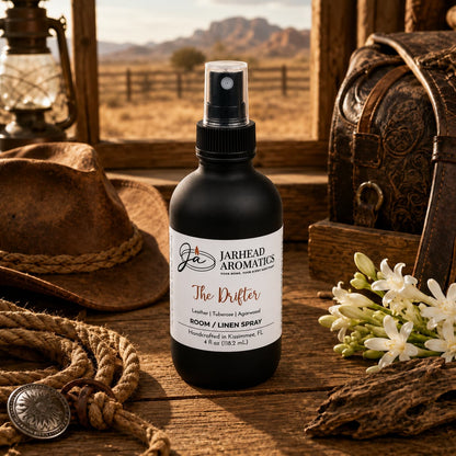 A bottle of Jarhead Aromatics The Drifter Room & Linen Spray | Leather, Tuberose & Smoky Agarwood rests on a wooden table alongside white flowers, a lantern, and a leather bag. With its western-inspired blend of leather, tuberose, and smoky agarwood, this fragrance feels right at home beside a rustic fence with distant mountains in the background.