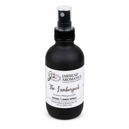 A black spray bottle labeled Jarhead Aromatics The Lumberjack Room & Linen Spray, featuring notes of mahogany, cedarwood, and oak, is handcrafted in Kissimmee, FL. This woodsy home fragrance comes with a black cap and is displayed against a clean white background.