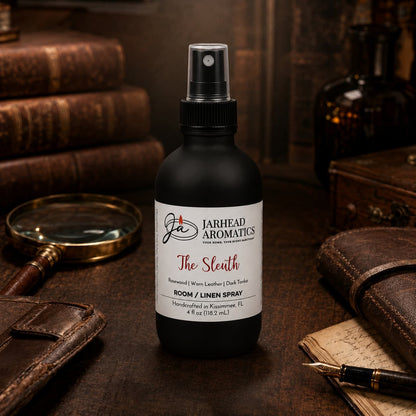 A bottle of Jarhead Aromatics’ The Sleuth Room & Linen Spray | Leather, Rosewood & Sandalwood rests on a desk alongside leather-bound books, a pen, and papers, infusing the scholarly vintage atmosphere with its sophisticated home fragrance featuring notes of leather, rosewood, and warm sandalwood.
