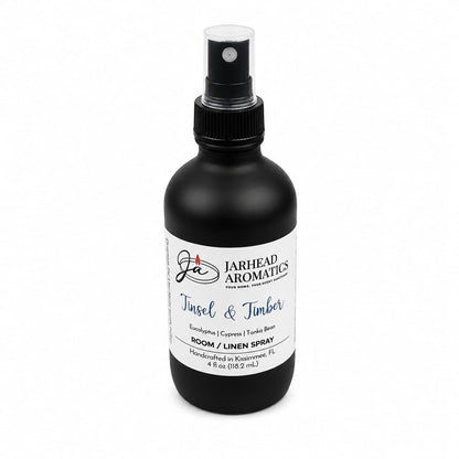 A black spray bottle with a white label reading Tinsel & Timber Room & Linen Spray, listing notes of eucalyptus, pine, smoke, and tonka bean for a winter forest scent.