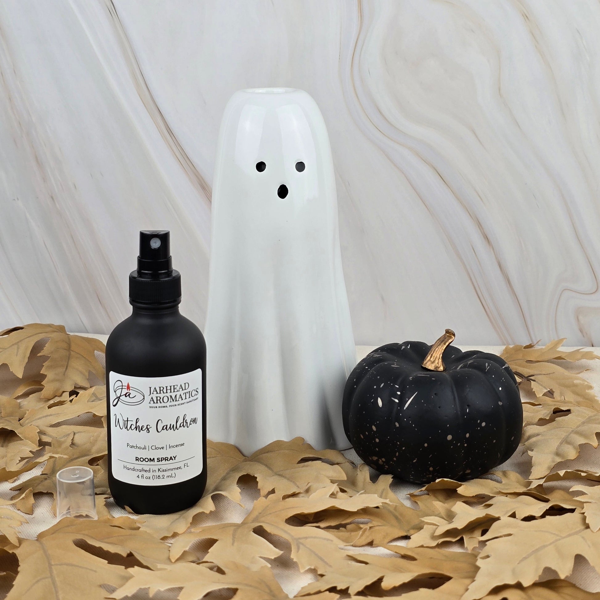 A white ceramic ghost, a black pumpkin, and Jarhead Aromatics’ Witches Cauldron Room Spray from the Halloween Collection are arranged on dried oak leaves against a marbled background.