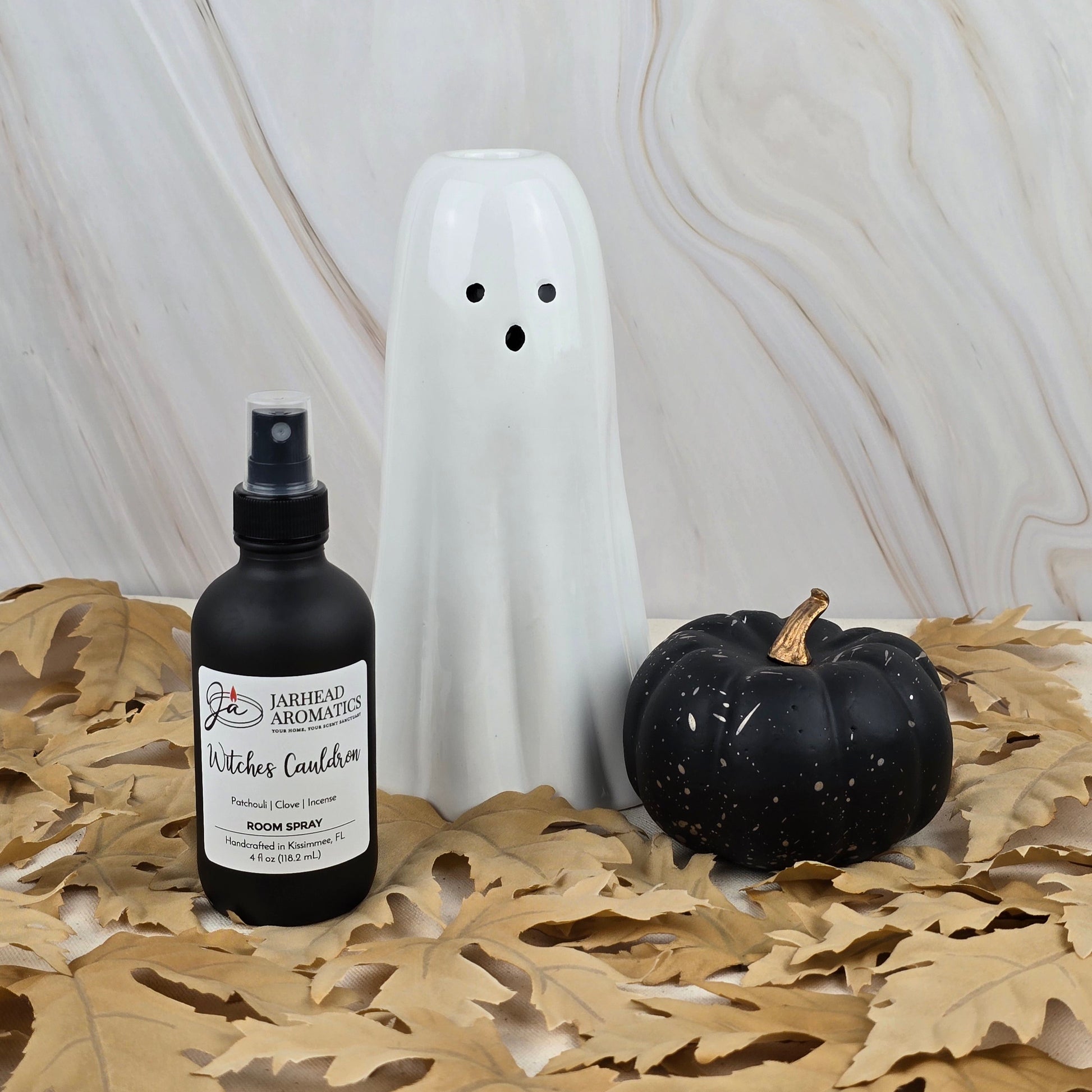 A white ceramic ghost, a black pumpkin, and Jarhead Aromatics Witches Cauldron Room Spray from the Halloween Collection rest on beige dried leaves against a light marble background.