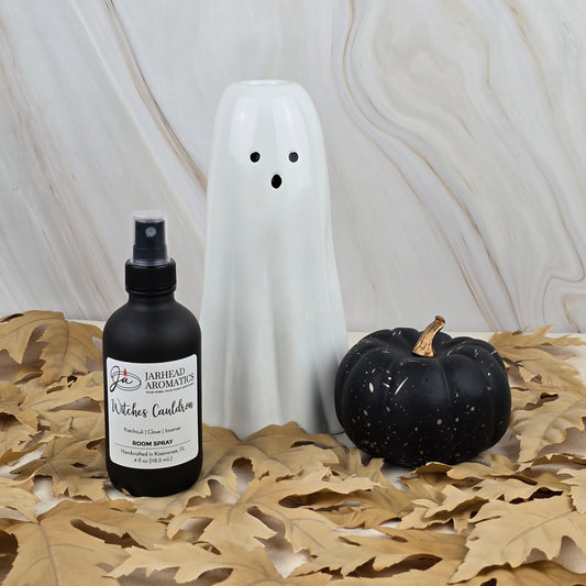 A white ceramic ghost, a black pumpkin, and Jarhead Aromatics Witches Cauldron Room Spray from the Halloween Collection rest on beige dried leaves against a light marble background.