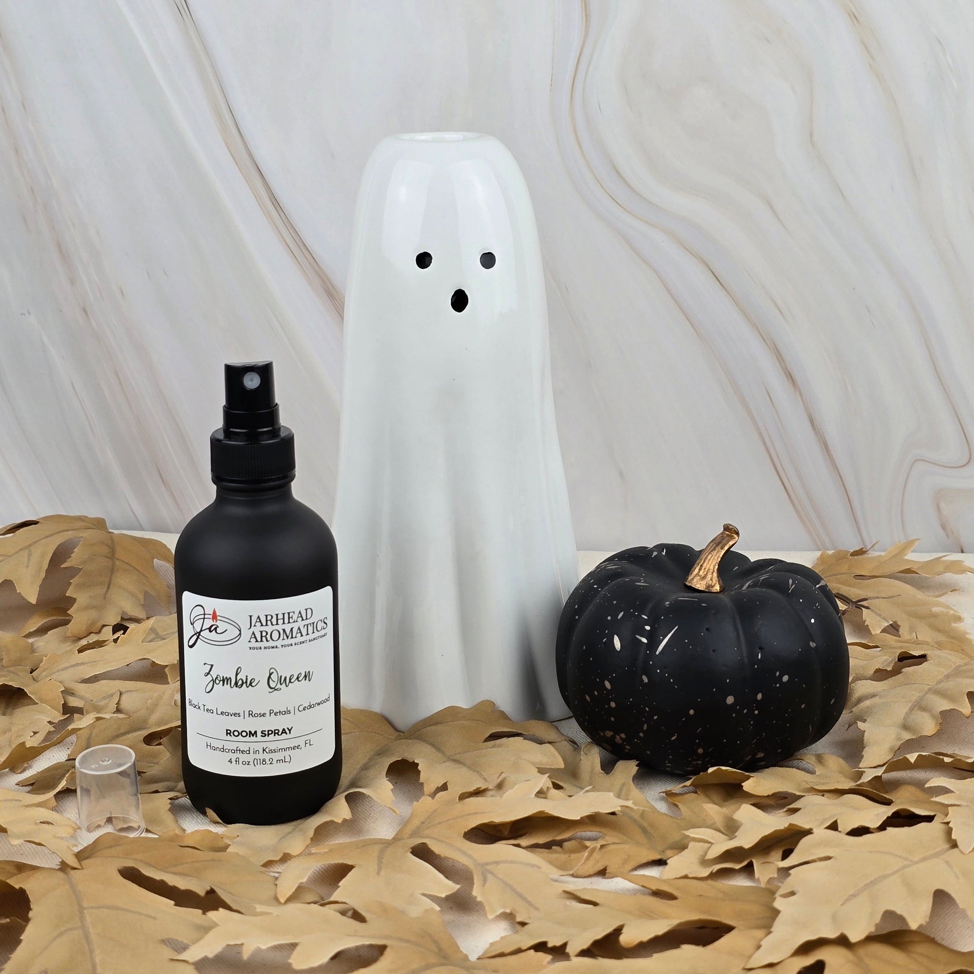 A white ceramic ghost, a black pumpkin, and Jarhead Aromatics Zombie Queen Room Spray from the Halloween Collection with gothic fragrance sit on beige leaves against a marble-patterned background.