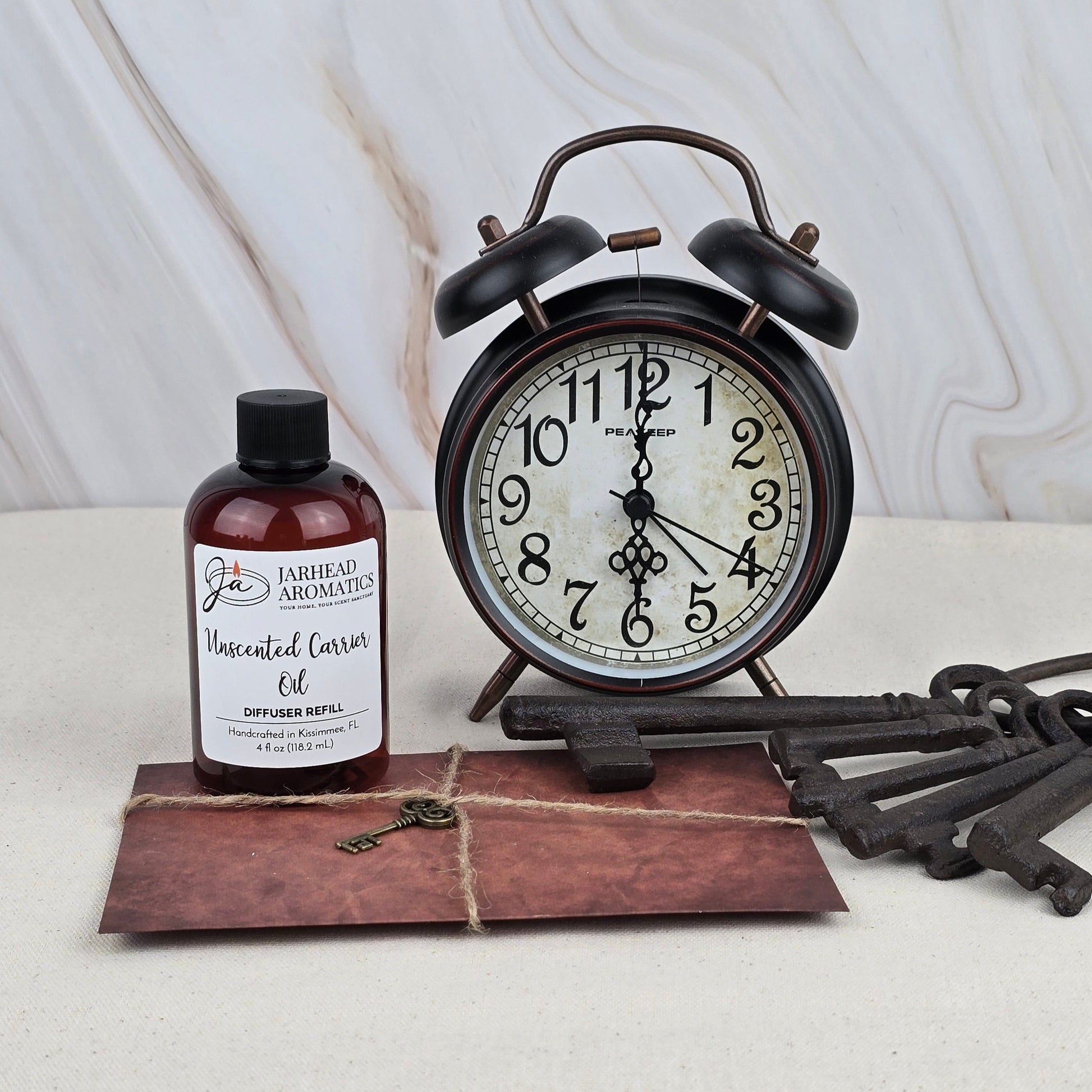 A vintage alarm clock, a bottle of Jarhead Aromatics Unscented Carrier Oil - Diffuser Refill, old-fashioned keys, and a tied brown envelope rest on marble, capturing the tranquility of a coastal retreat.