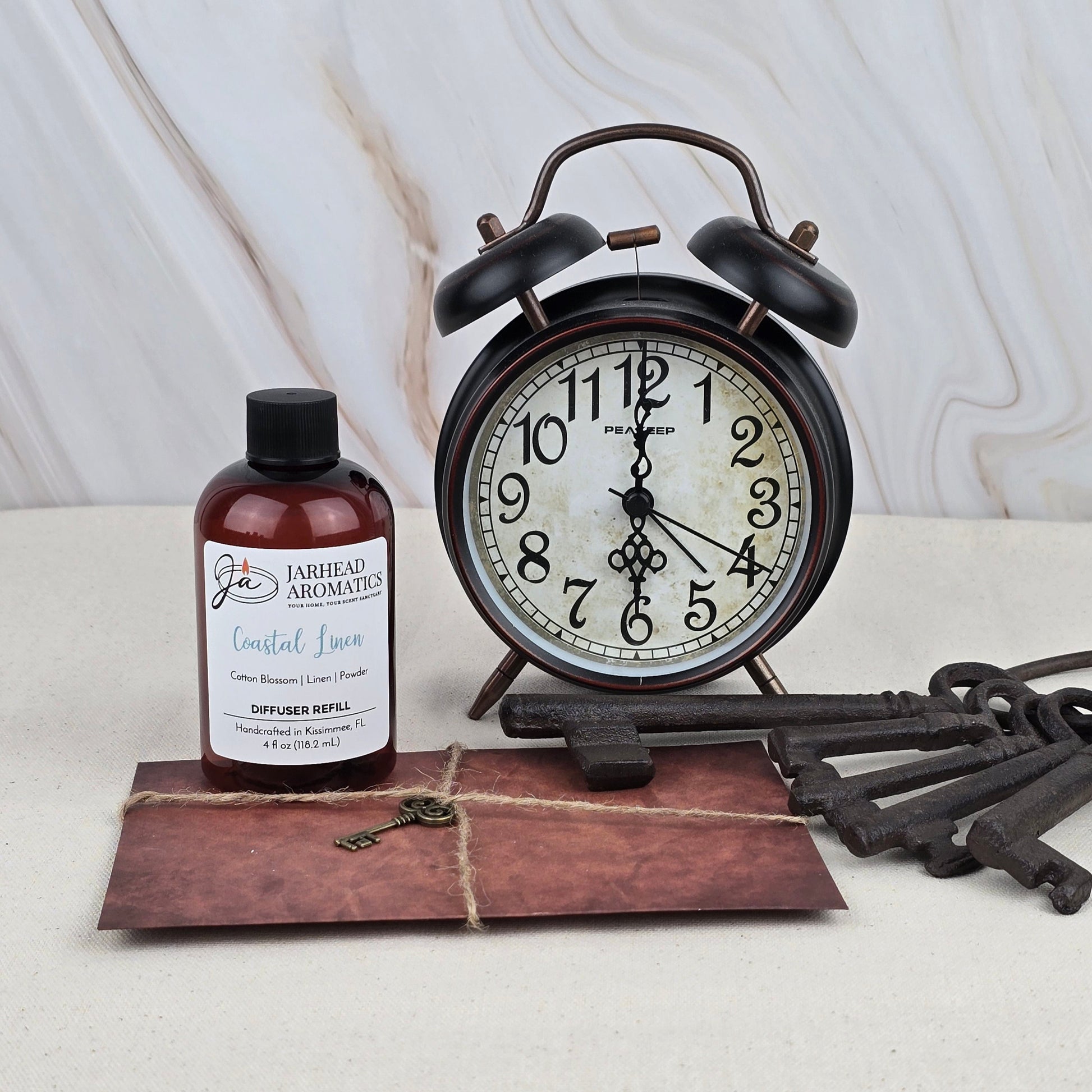 A vintage black alarm clock, an old-fashioned key, a brown envelope tied with twine, and a bottle of Jarhead Aromatics Coastal Linen - Diffuser Refill are arranged on a light surface against a marbled background.