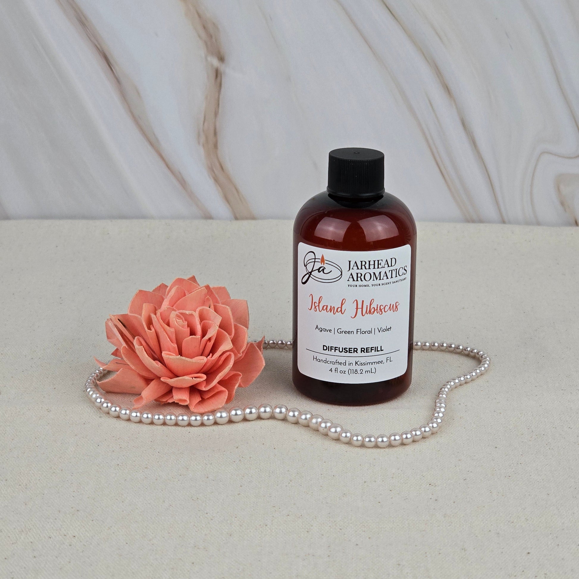 A bottle of Jarhead Aromatics Island Hibiscus - Diffuser Refill sits on a bright surface beside a pink artificial flower and pearls, capturing tropical vibes against a marble-style background.