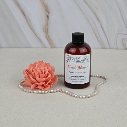 A bottle of Jarhead Aromatics Island Hibiscus - Diffuser Refill sits on a bright surface beside a pink artificial flower and pearls, capturing tropical vibes against a marble-style background.