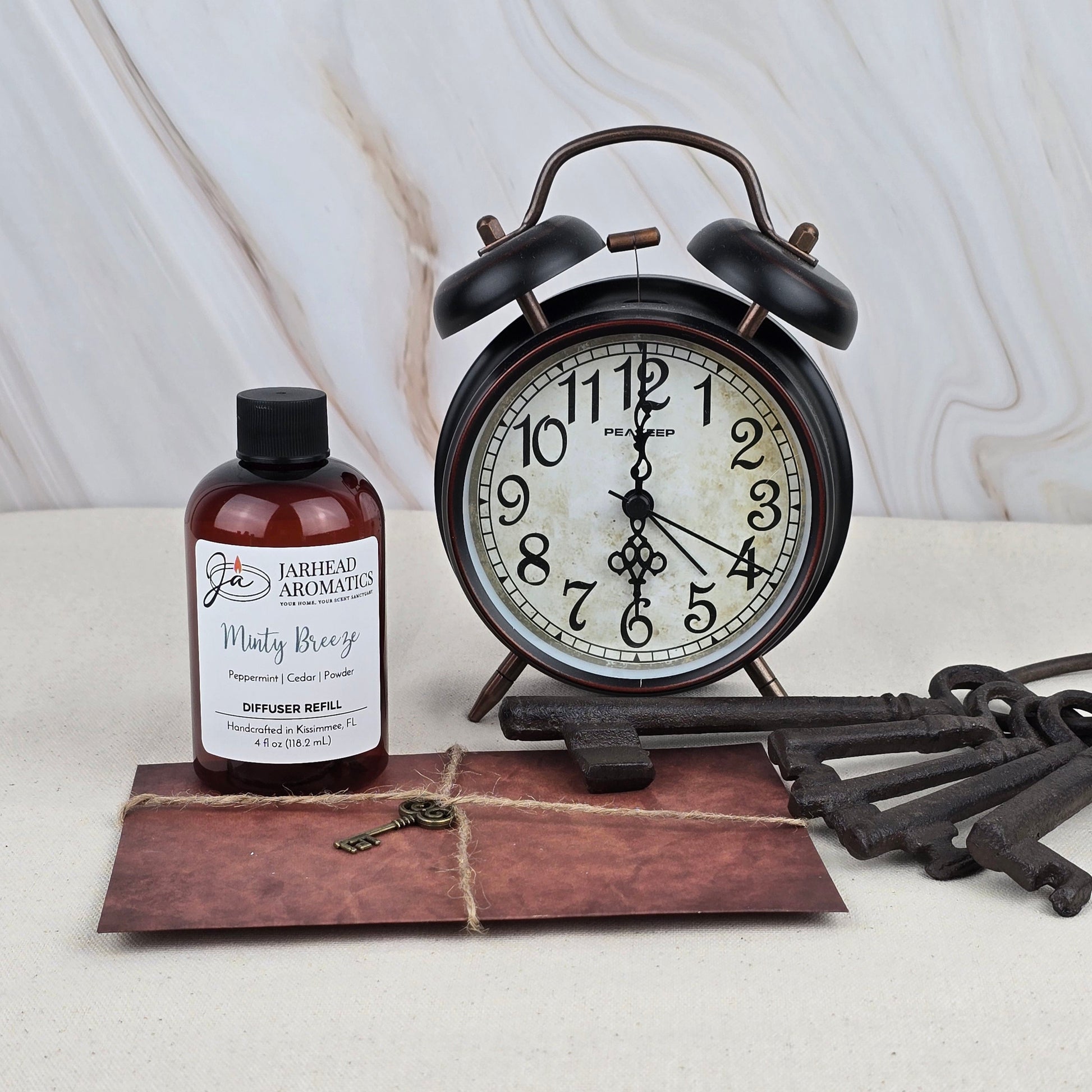 A vintage black alarm clock, large antique keys, a brown twine-tied envelope, and a Jarhead Aromatics Minty Breeze Diffuser Refill are arranged on a light surface with a marble background.