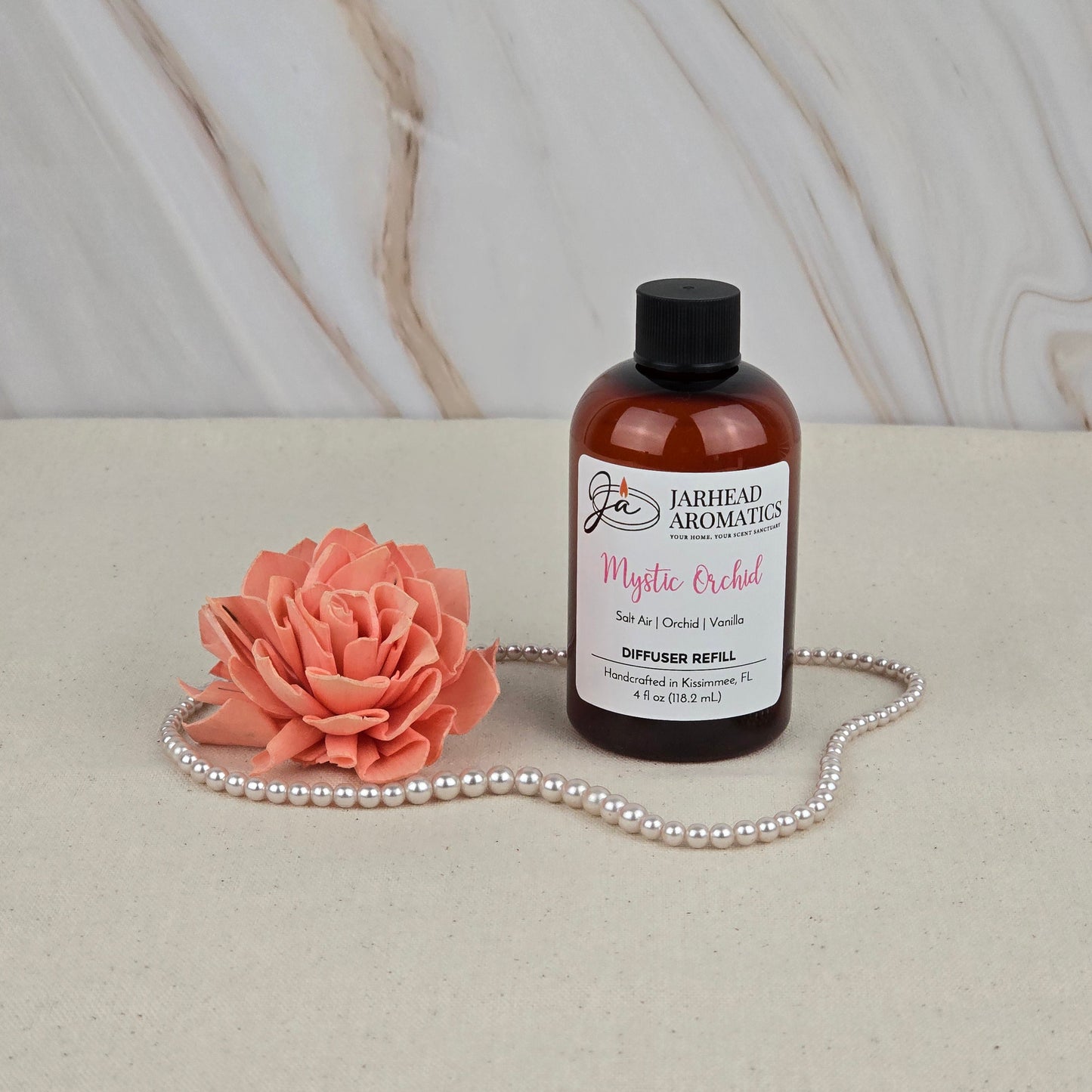 A brown bottle of Jarhead Aromatics Mystic Orchid - Diffuser Refill rests on a beige surface beside a peach artificial flower and pearls, bringing a coastal freshness to the scene against a light marble-patterned background.