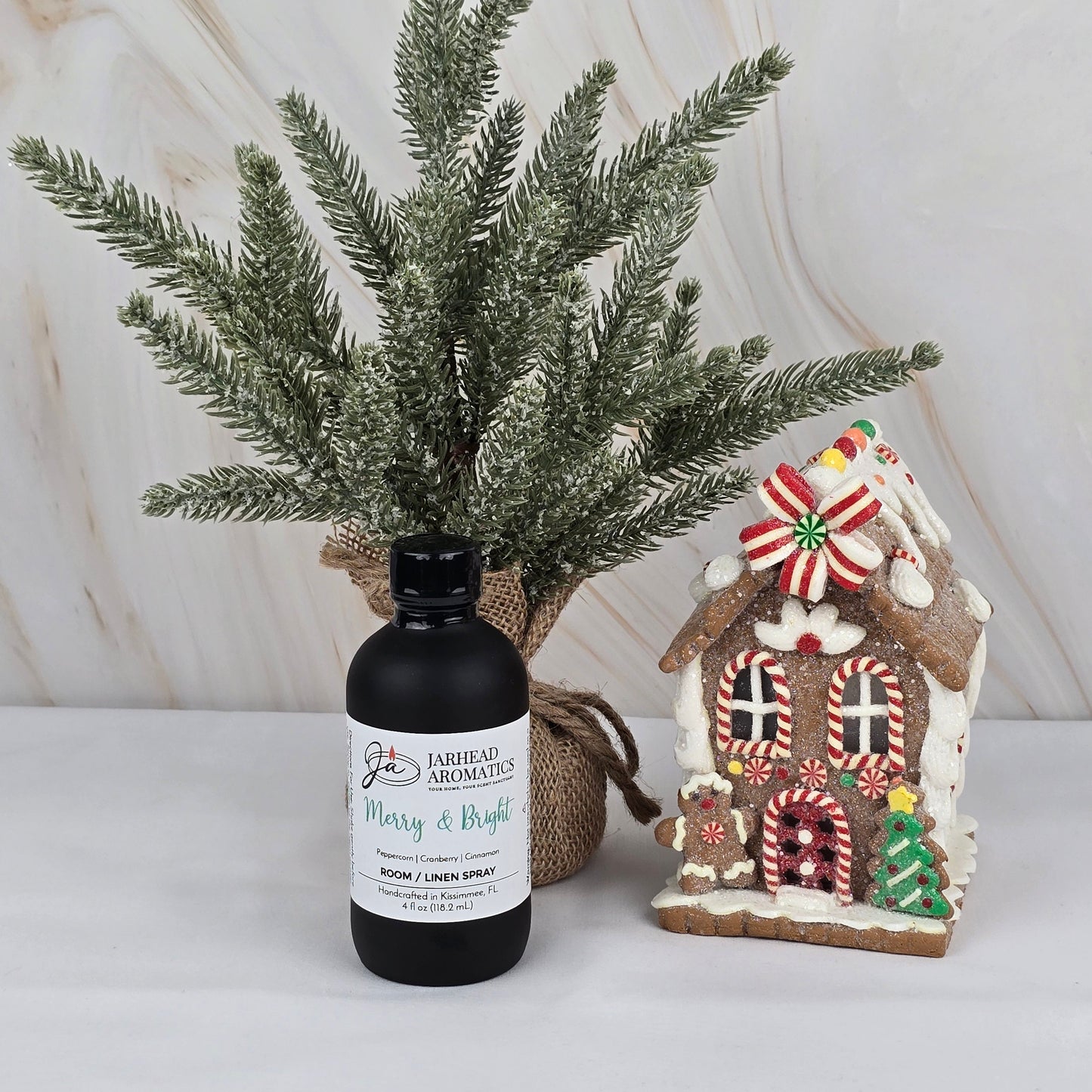 A bottle of Jarhead Aromatics’ “Merry & Bright” Room/Linen Spray from the Winter Collection rests on a white surface beside a small frosted pine in burlap and a decorative gingerbread house with candy accents.